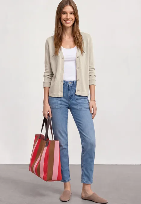 Frau trägt Cardigan, weißes Top, blaue Jeans, bequeme Schuhe und bunte gestreifte Tragetasche.