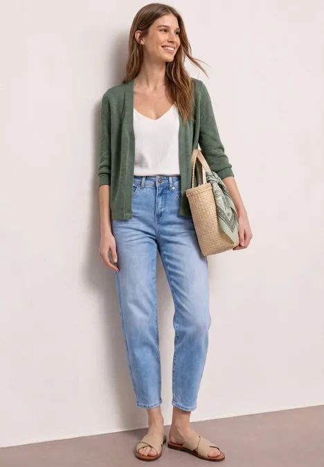 Vrouw leunt tegen muur, witte top, groene cardigan, lichte jeans, sandalen en rieten tas