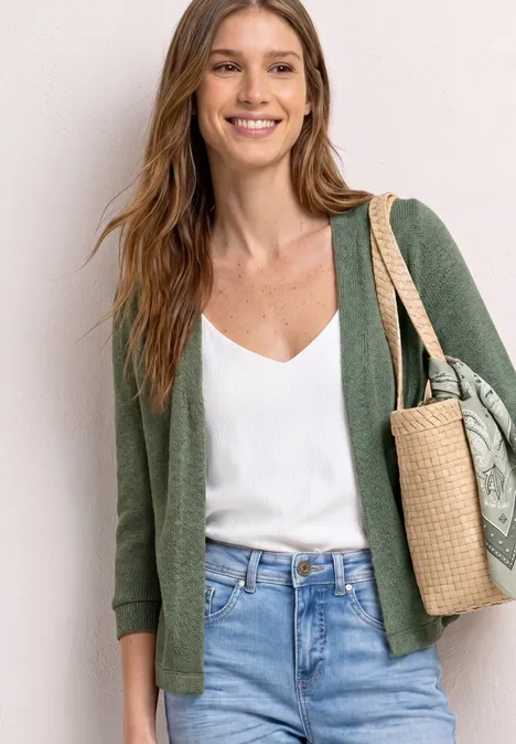 Vrouw in een groen vest, wit topje en jeans met een gevlochten strooien tas over haar schouder