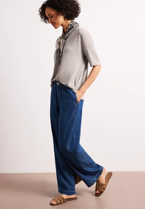 Frau im Profil trägt graues Shirt, weite blaue Jeans, Ledersandalen und Seidenschal.