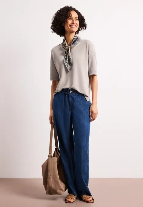 Frau vor weißem Hintergrund, trägt beiges Oberteil, weite blaue Hose, Halstuch und Tasche