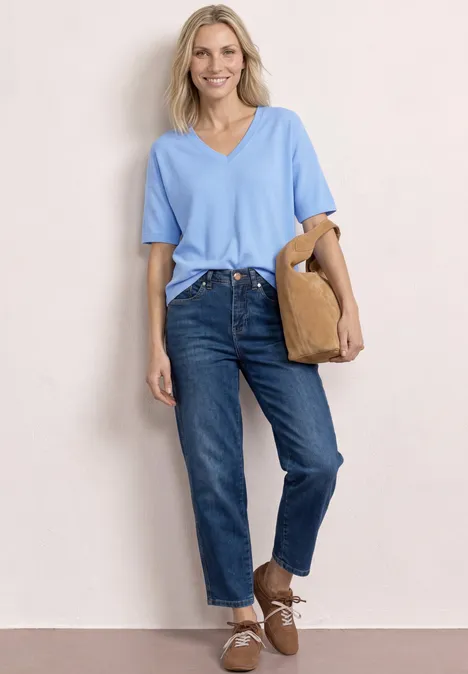 Frau mit hellblauem V-Shirt, blauen Jeans und braunen Schuhen lehnt an der Wand und trägt eine beige Tasche.