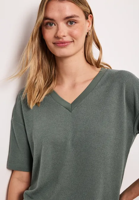 Frau im khakigrünen V-Shirt, kurzärmelig, vor hellem Hintergrund.