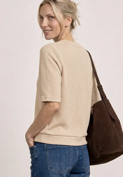 Frau im beigen Kurzarm-Pullover, blauer Jeanshose und brauner Umhängetasche, seitlich stehend.