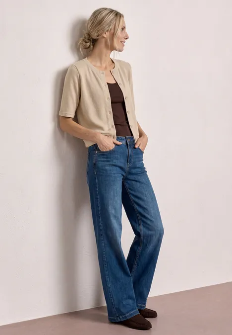 Model trägt beige Kurzarm-Cardigan, braunes Top und weite Jeans, steht seitlich an Wand.