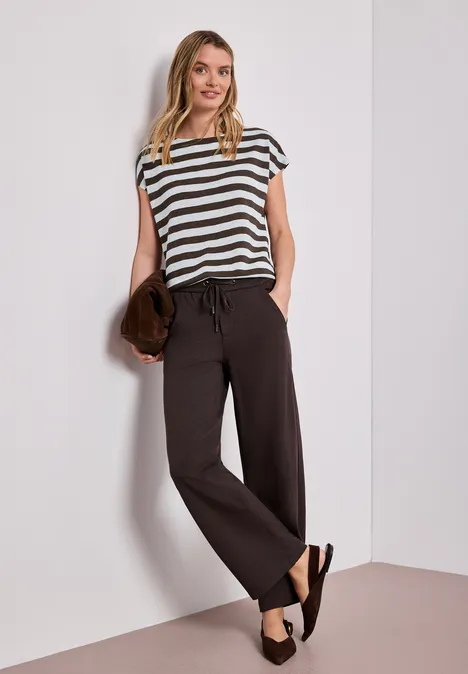 Vrouw leunt tegen muur, draagt gestreept t-shirt, wijde zwarte broek, sandalen en clutch in hand.