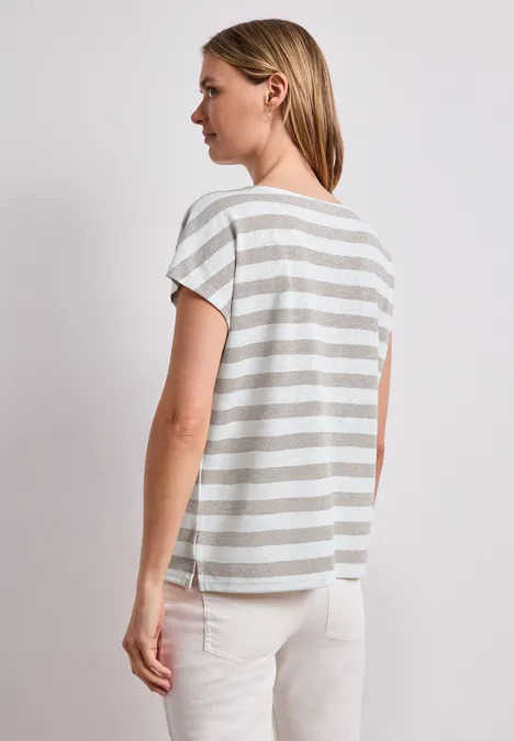 Vrouw van opzij met de rug naar de camera, draagt een grijs-wit gestreept T-shirt en een lichte broek.