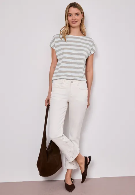 Vrouw in gestreept T-shirt, witte jeans, bruine tas en schoenen, voor een witte achtergrond.