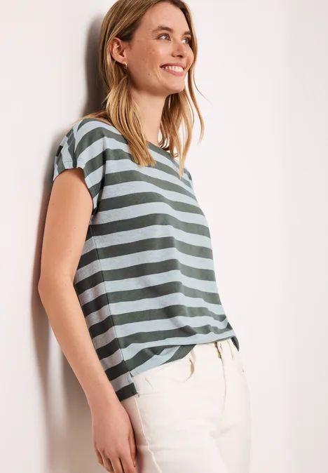 Eine Frau lehnt an einer Wand, trägt ein kurzärmeliges, grün-grau gestreiftes T-Shirt und helle Hosen.