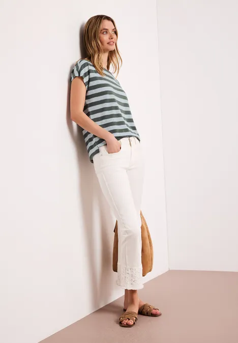 Frau lehnt an Wand, trägt grün-weiß gestreiftes T-Shirt, weiße Schlaghose mit Fransen und Sandalen.