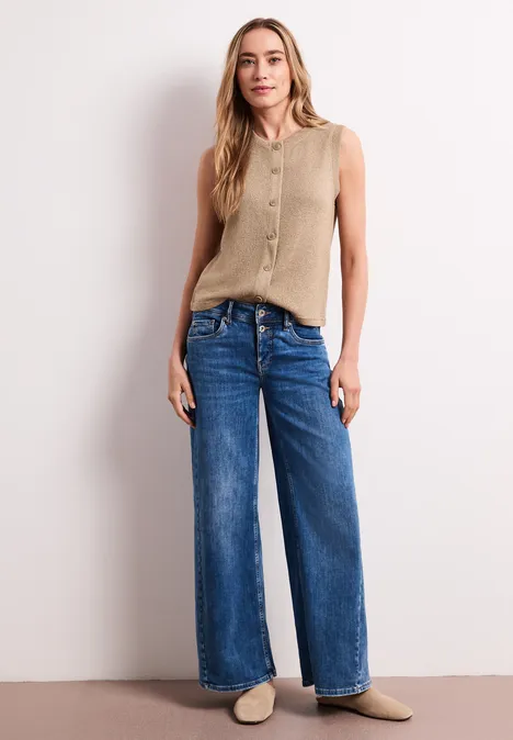 Frau in beigem Tanktop mit Knopfleiste, weitem blauen Jeans und hellen Hausschuhen vor weißer Wand.