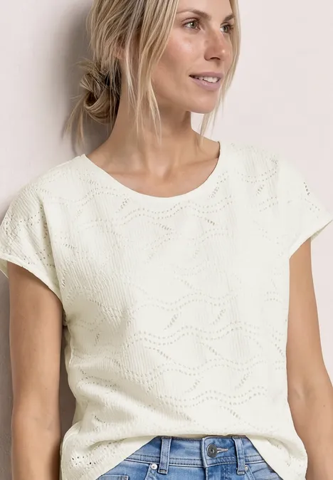 Frau in weißem, kurzärmeligem Strickoberteil mit Wellenmuster, kombiniert mit Jeans.