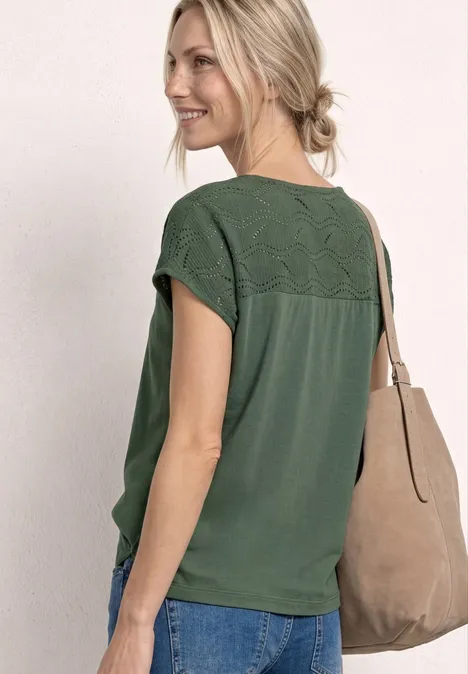 Frau trägt grünes Spitzenshirt, blaue Jeans und beige Schultertasche, das Haar zum lockeren Dutt gebunden.