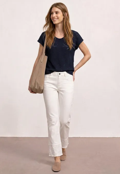 Eine Frau in schwarzem T-Shirt, weißen Jeans und beigen Mules trägt eine große Tasche vor hellem Hintergrund.