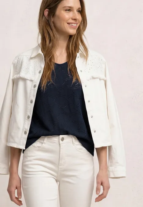 Frau trägt weiße Jeansjacke, weiße Hose und dunkles Top vor neutralem Hintergrund.