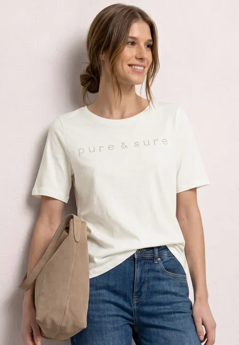 Frau in weißem T-Shirt mit 'pure&süre'-Print, blauen Jeans und beiger Schultertasche.