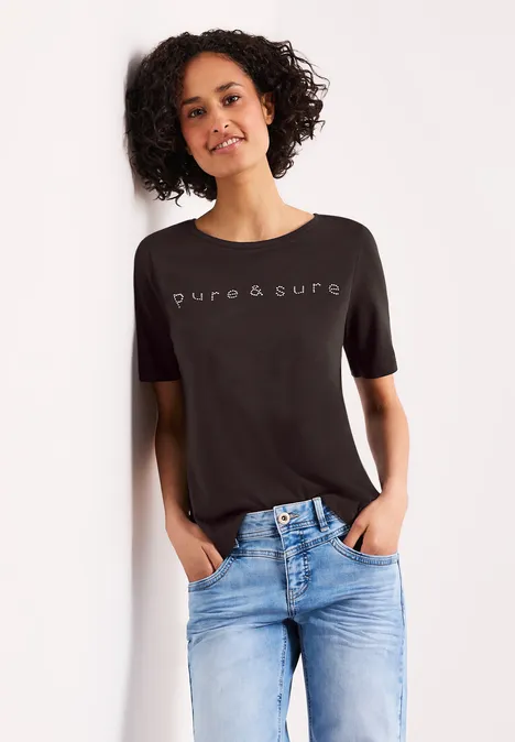Vrouw leunt tegen muur, draagt een zwart 'pure&sure' T-shirt en een blauwe jeans.