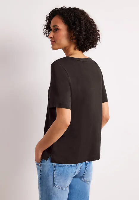 Vrouw met krullend haar draagt een zwart T-shirt en blauwe jeans, staand in zijaanzicht met handen in zakken.