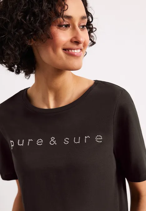 Een lachende vrouw draagt een zwart T-shirt met de tekst "pure & sure" en kijkt opzij.