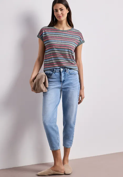 Frau in gestreiftem T-Shirt, hellblauen Jeans, trägt hellen Pullover und bequeme Slipper vor neutralem Hintergrund.