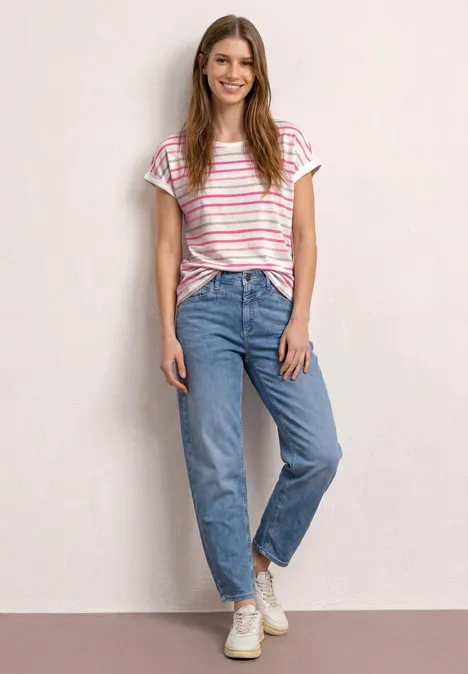 Een vrouw in een gestreept t-shirt, lichtblauwe jeans en witte sneakers staat voor een lichte muur.