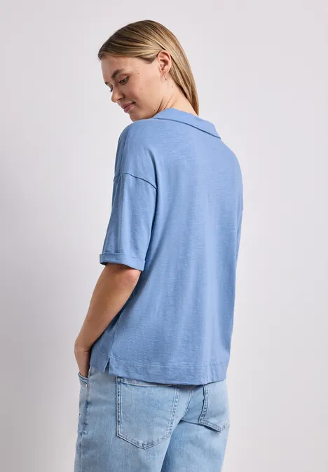 Frau von hinten in lockerem hellblauem Kurzarmshirt und Jeans vor weißem Hintergrund.