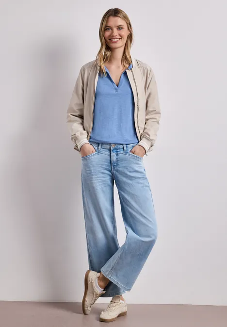 Person in blauem Shirt, beiger Jacke, weiten Jeans und Sneakers steht lässig mit Händen in den Taschen.