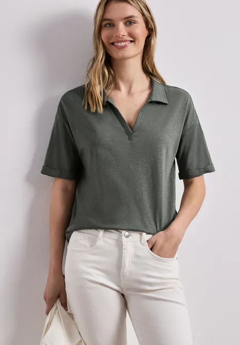 Frau trägt olivgrünes, kurzärmeliges Poloshirt und helle, hochgeschnittene Jeans, hält Tasche.