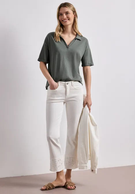 Frau in dunkelgrünem Shirt, weiße Culotte mit Spitzenbesatz, Sandalen, hält einen Blazer.