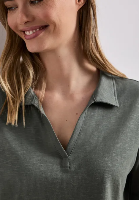 Eine Person mit langen Haaren trägt ein grünes Poloshirt mit offenem Kragen.