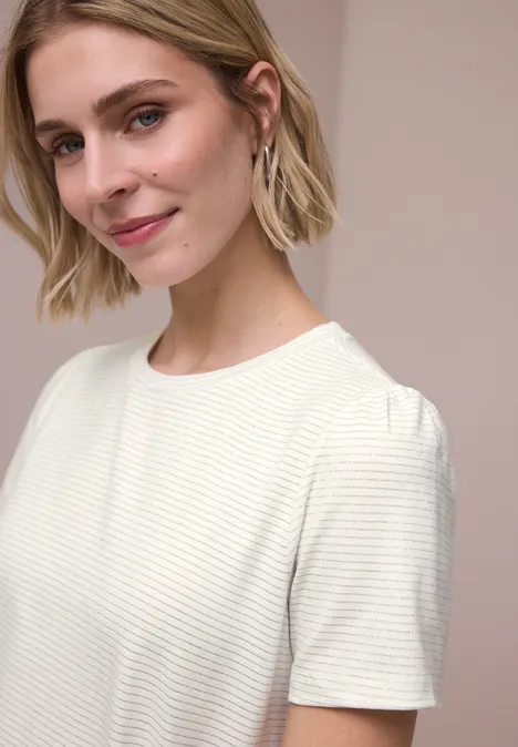 Frau mit kurzem blonden Haar, trägt ein weißes geripptes Kurzarmshirt vor neutralem Hintergrund.