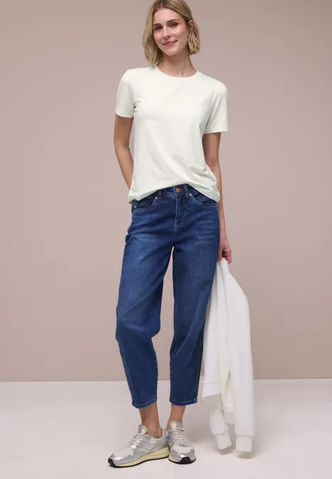 Person in weißem T-Shirt, blauen High-Waist-Jeans und Sneakers, hält weiße Jacke vor schlichtem Hintergrund.