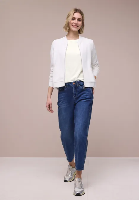 Eine Frau trägt eine weiße Jacke, helles Shirt, blaue Jeans und silberne Schuhe vor beigem Hintergrund.