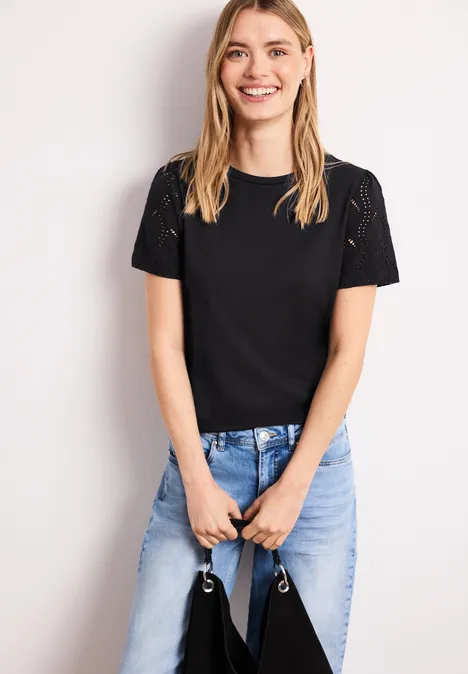 Frau mit schwarzem T-Shirt und hellblauen Jeans steht und hält eine Tasche vor dem Körper.