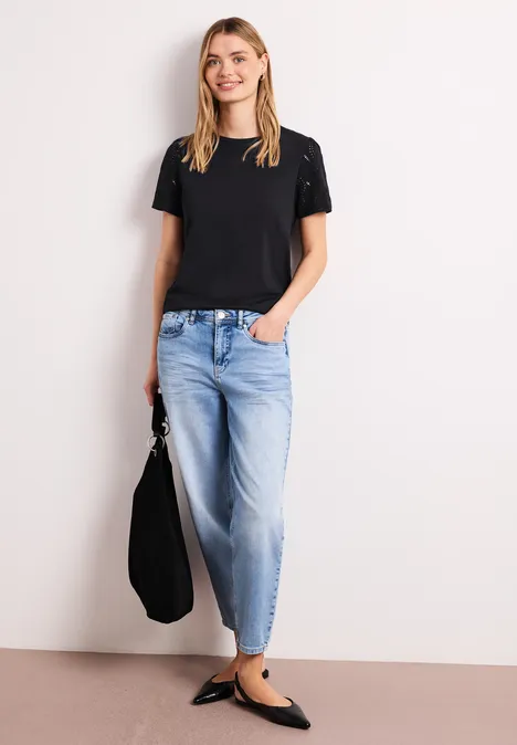 Person vor hellem Hintergrund mit schwarzem T-Shirt, weiten Jeans, schwarzen Ballerinas und Tasche.