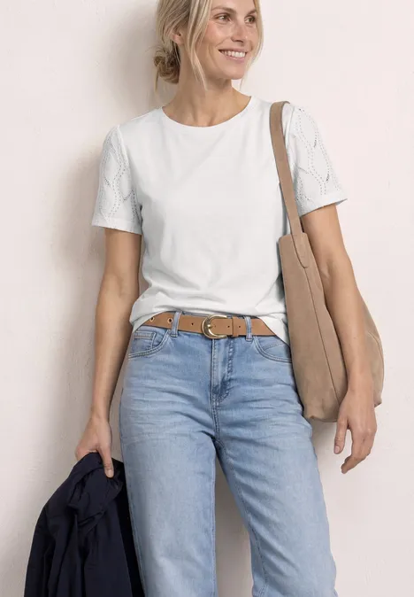 Person in weißem T-Shirt, blauen Jeans und Gürtel, trägt eine Schultertasche und hält eine Jacke in der Hand.