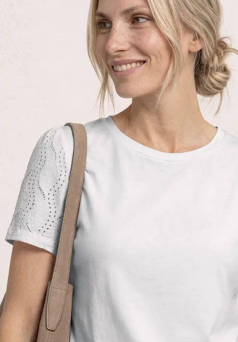 Lächelnde Frau in weißem T-Shirt mit Lochstickärmeln und Schultertasche vor hellem Hintergrund.