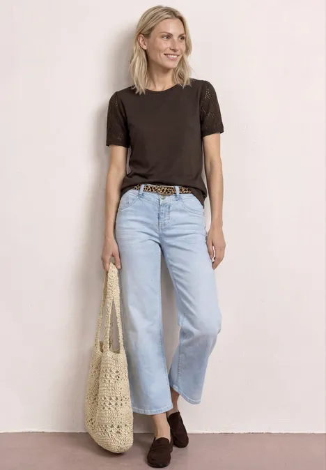 Frau in schwarzem T-Shirt, hellblauer, weiten Jeans, braunem Gürtel und Häkeltasche vor hellem Hintergrund.