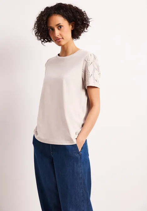 Frau trägt cremefarbenes T-Shirt mit Spitzenärmeln und weiten blauen Jeans, Hände in den Taschen.