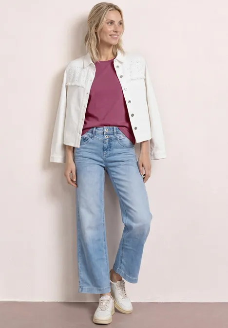 Een vrouw in lichtblauwe jeans, bordeauxrood shirt, witte jas en sneakers voor een roze achtergrond.