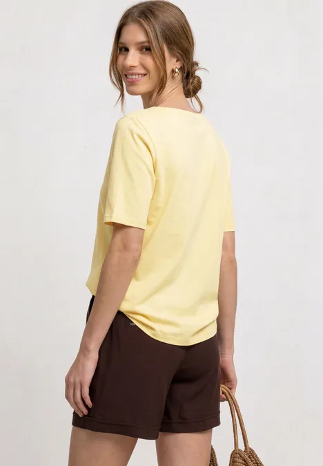 Frau mit gelbem T-Shirt und braunen Shorts von hinten, trägt Strohkorb und blickt zur Seite.