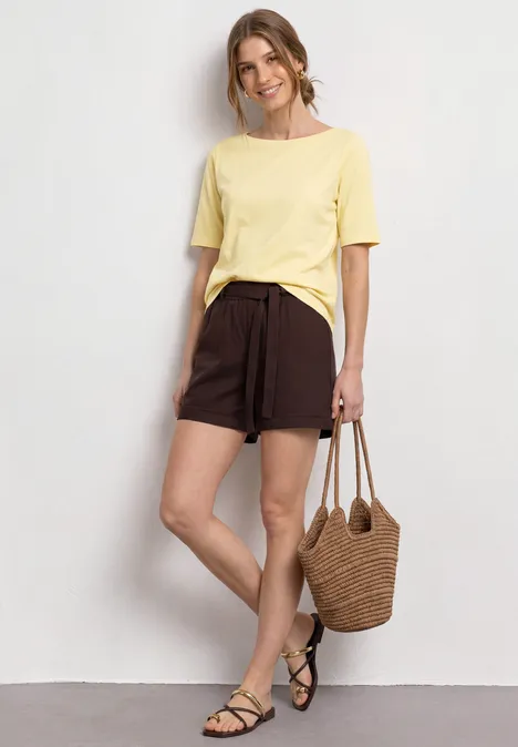 Person in gelbem T-Shirt, braunen Shorts, mit Basttasche und schwarzen Sandalen an Wand.