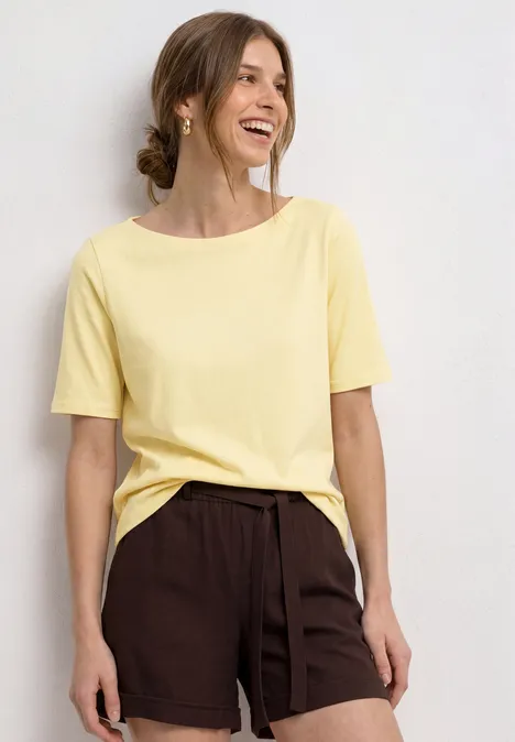 Frau in hellem T-Shirt und braunen Shorts vor weißem Hintergrund.