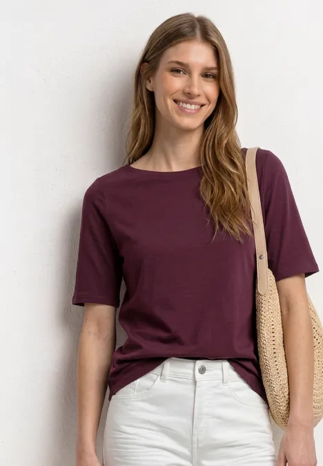 Eine Frau trägt ein dunkelviolettes T-Shirt, weiße Hose und eine Strohtasche.