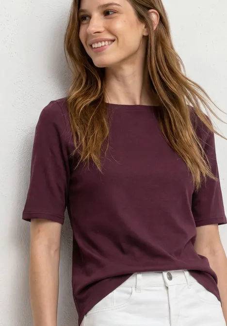 Frau in bordeauxfarbenem T-Shirt und hellbeiger Hose vor weißer Wand.