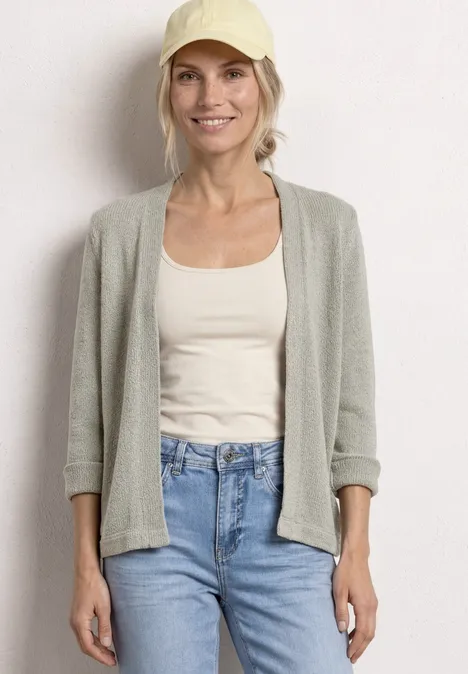 Frau in grauem Cardigan, weißem Top und Jeans steht vor hellem Hintergrund.