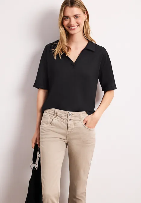 Eine Frau trägt eine schwarze V-Ausschnitt-Bluse und beigefarbene Hose, hält eine kleine schwarze Handtasche.