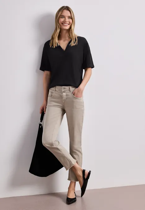 Frau in schwarzem Oberteil und beigen Hosen mit schwarzer Tasche lehnt an weißer Wand.