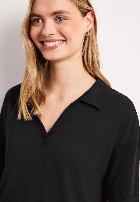 Lächelnde blonde Frau im schwarzen Hemd mit V-Ausschnitt und Kragen vor hellem Hintergrund.