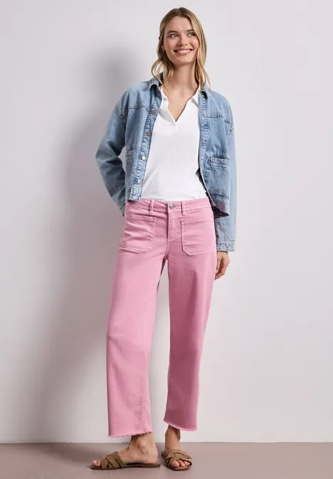 Eine Frau steht vor neutralem Hintergrund, trägt eine Jeansjacke, ein weißes Oberteil und rosafarbene, weite Hosen.